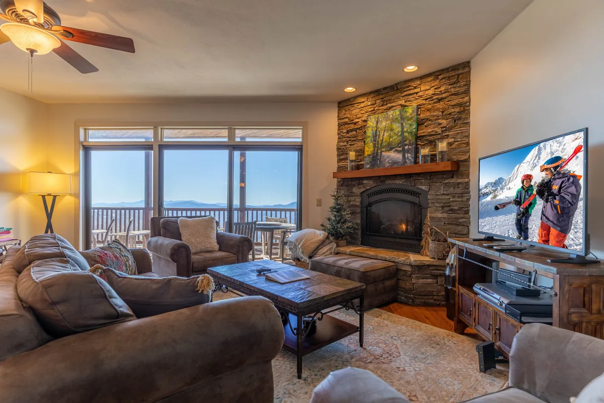 The image features a cozy living area with plush seating arranged around a crackling fireplace. Expansive windows frame breathtaking mountain vistas, inviting in natural light and offering panoramic sunset views. A TV is positioned for entertainment, and the ambiance is warm, inviting guests to unwind after a day of outdoor adventure.