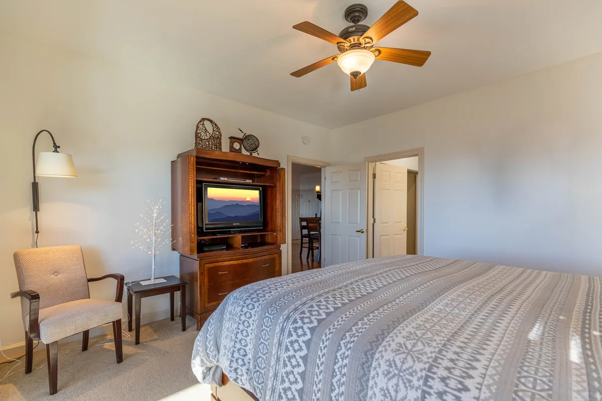 A cozy queen bedroom with a patterned bedspread, wooden entertainment unit, accent chair, and ceiling fan. The space feels restful and welcoming, offering a comfortable hideaway for guests or family, enhanced by thoughtful decor and natural light.