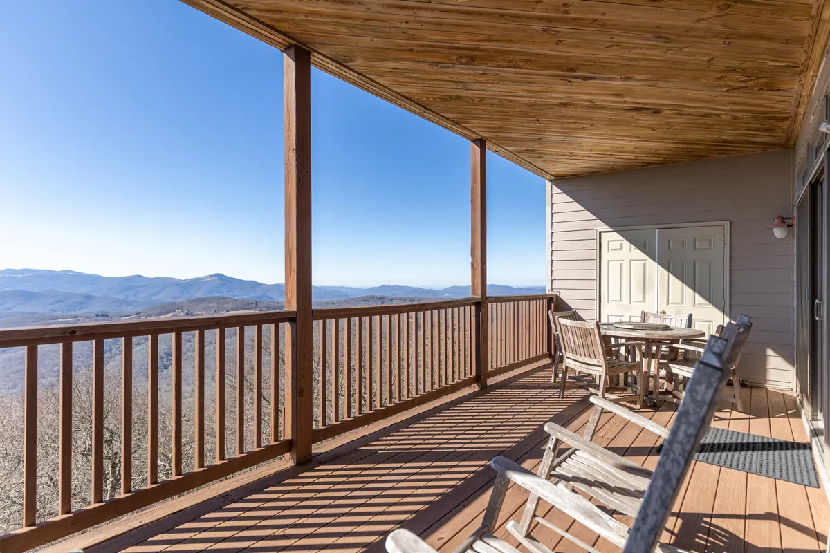 A covered deck with wooden railings overlooks a breathtaking mountain panorama. Rocking chairs and a table invite guests to relax, sip coffee, or watch the sun rise or set over the peaks. The space blurs the line between indoors and outdoors, offering tranquility and stunning views.