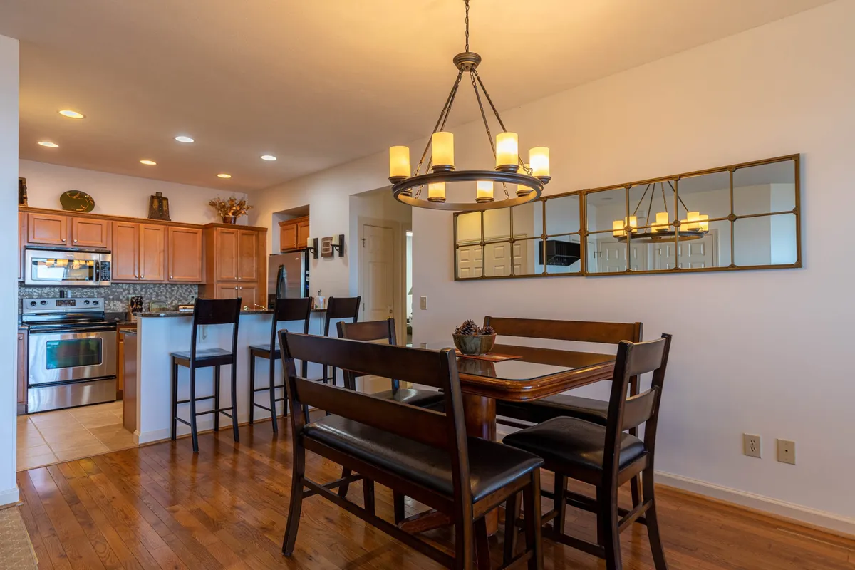 An inviting dining area with a table set for six, comfortable chairs, and a warm chandelier overhead. The kitchen counter is visible in the background, establishing a connection between cooking and dining spaces and suggesting a seamless flow for group meals and gatherings.
