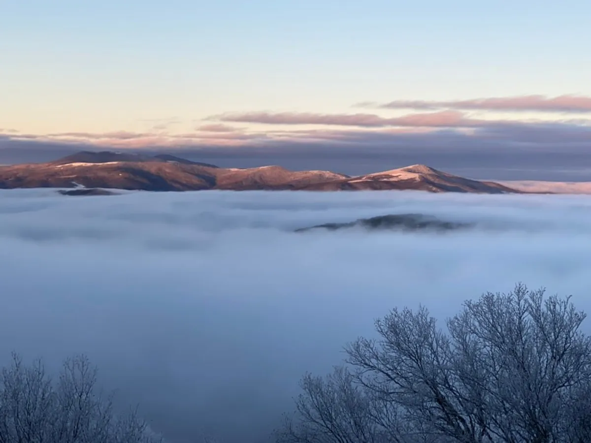 A breathtaking view image featuring towering mountain peaks, a fog-covered valley, and frosted trees. This tranquil scene invites exploration, with visible hiking trails and opportunities for nature photography or meditative walks. The sunrise light and fog rolling over the mountains set a magical, early-morning tone.