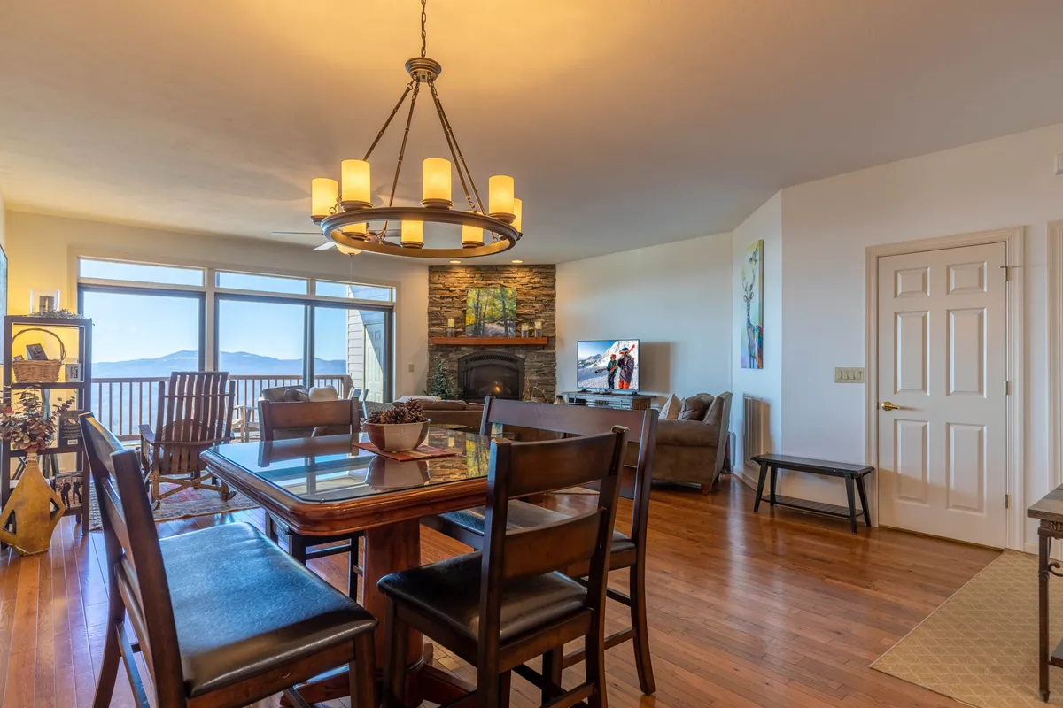 A spacious, inviting dining area centered around a wooden table, with a glowing chandelier overhead and a fireplace off to the side. Large windows frame scenic views and allow natural light to fill the room, suggesting a strong connection to the outdoors and a perfect spot for sunset toasts or intimate dinners.