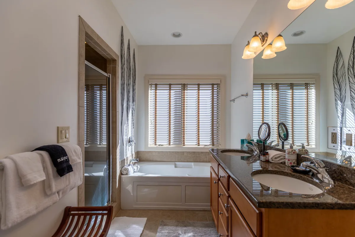 A spacious, spa-inspired bathroom features a deep soaking bathtub, double vanity sinks, a large window with blinds for natural light, and a beautifully tiled shower area. Decorative wall art and soft lighting create an inviting, calming ambiance ideal for unwinding.