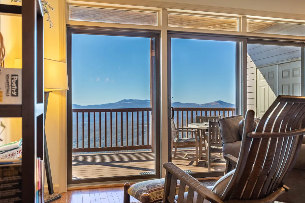 A cozy interior setting where large windows overlook the mountains, a comfortable armchair is positioned to take in the view, and a small table sits on the adjacent balcony. The space feels relaxed and inviting, blending the comforts of resort living with the beauty of the outdoors.