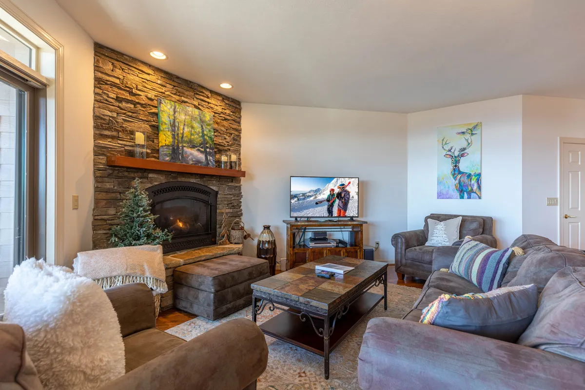 A welcoming interior scene featuring a stone fireplace, comfortable seating, a television, and thoughtful decor. The arrangement fosters socializing and relaxation, ideal for family gatherings by the fire with mountain vistas visible through windows.
